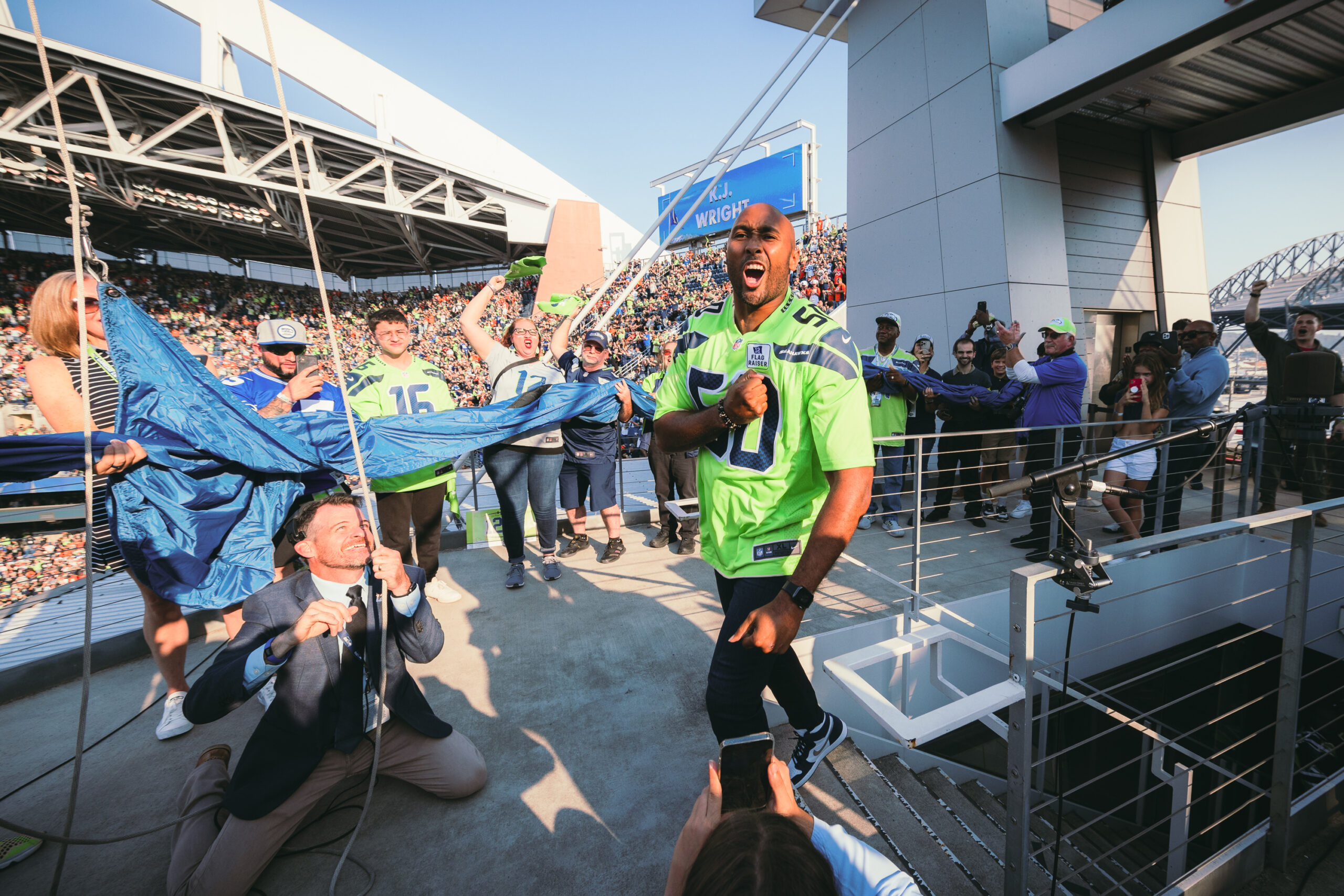 A man at Seattle's Lumen Field prepares to raise a blue flag with a white "12," a crowd is gathered behind him.