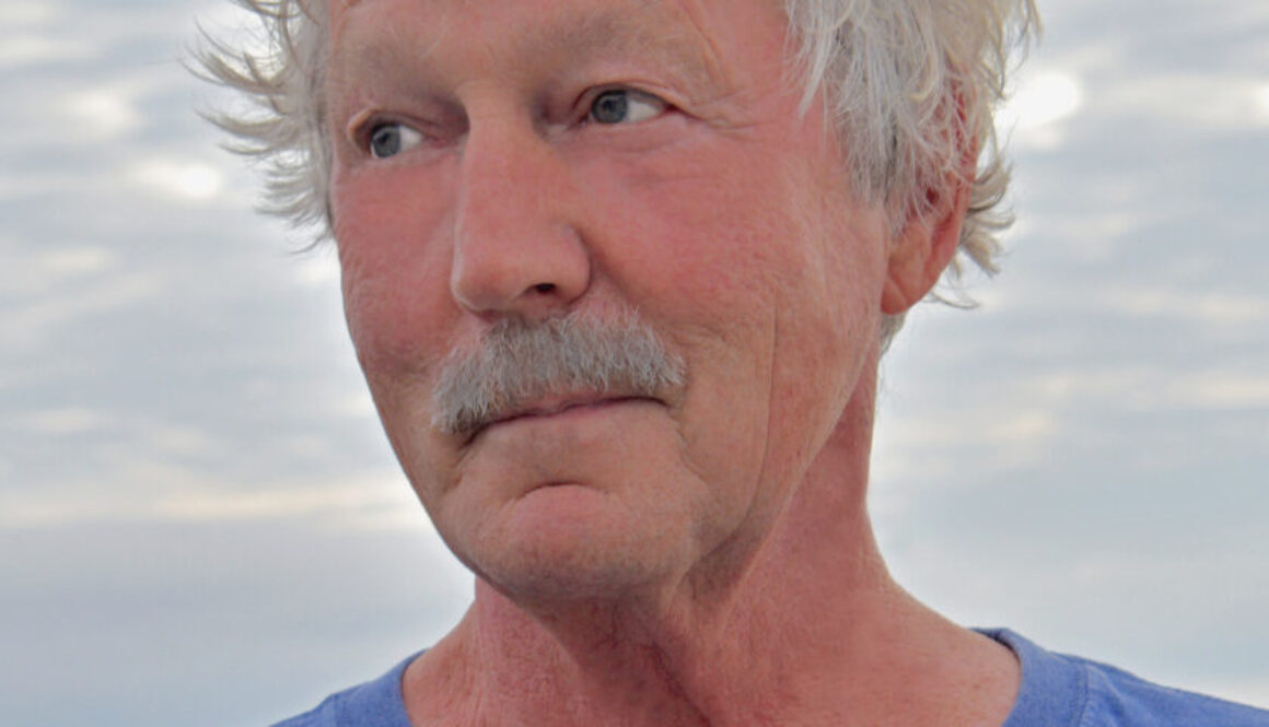 Portrait of Tim Pilgrim, looking off frame, in a blue shirt with a cloudy background.