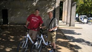 Jeff Kramer (left) and Mitch Evich stnad holding the handlebars of bikes in the shade of a tree.
