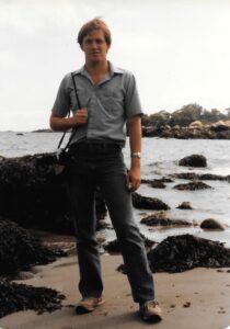 A man stands in front of a bay with the tide pooling behind him. A camera is slung over his shoulder and he looks at the camera.