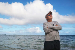 A casually dressed man wearing sunglasses, a baseball cap, and a long-sleeved shirt stands with his arms crossed, looking upward. He’s positioned in front of a calm, expansive body of water under a mostly blue sky with white clouds.