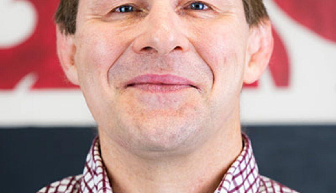 A man with blue eyes and brown hair smiles. He wears a red and white checkered shirt.