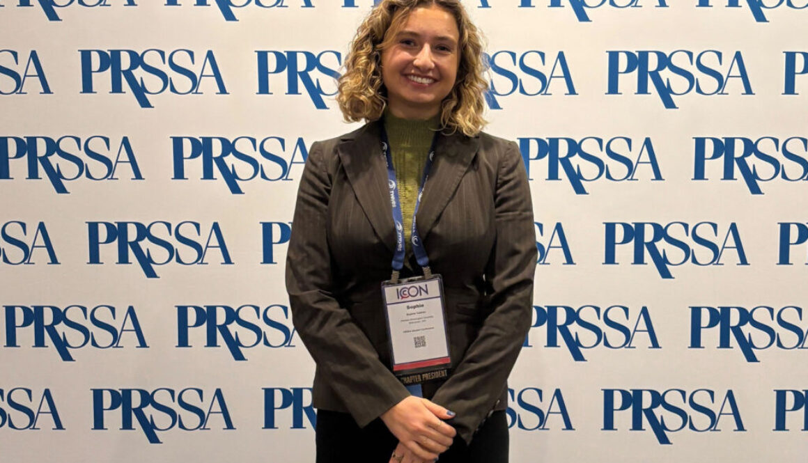 A woman in a business suit and badge stands smiling before a PRSSA branded backdrop.