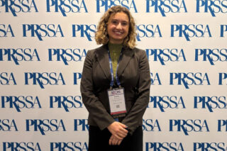 A woman in a business suit and badge stands smiling before a PRSSA branded backdrop.