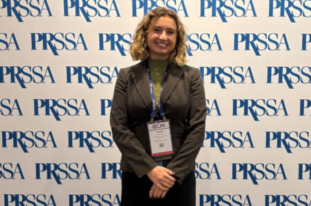 A woman in a business suit and badge stands smiling before a PRSSA branded backdrop.