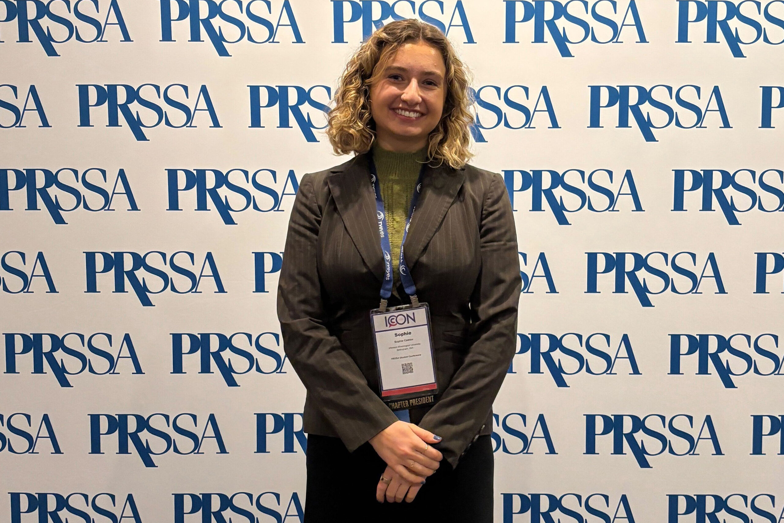 A woman in a business suit and badge stands smiling before a PRSSA branded backdrop.