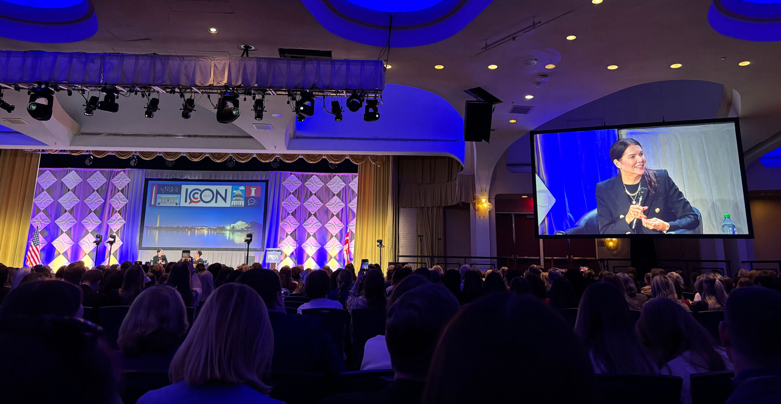 A conference room with a speaker on stage, shown on a large screen, and a captivated audience.