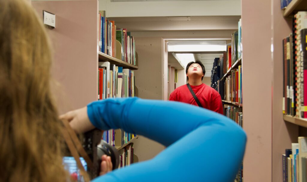 A student takes a photo of another student between bookshelves in a library.