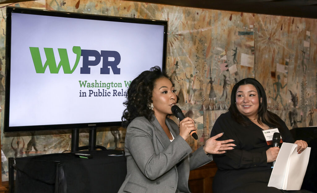 A woman presents in front of a TV screen while another woman listens.